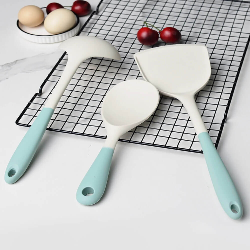 A set of three kitchen utensil set silicone tools, including a ladle, a serving spoon, and a spatula, displayed on a black cooling rack. The utensils have white silicone heads with ergonomic pastel green handles featuring hanging holes for convenient storage. Fresh eggs and cherries are placed nearby on a white countertop, enhancing the modern and elegant kitchen aesthetic.