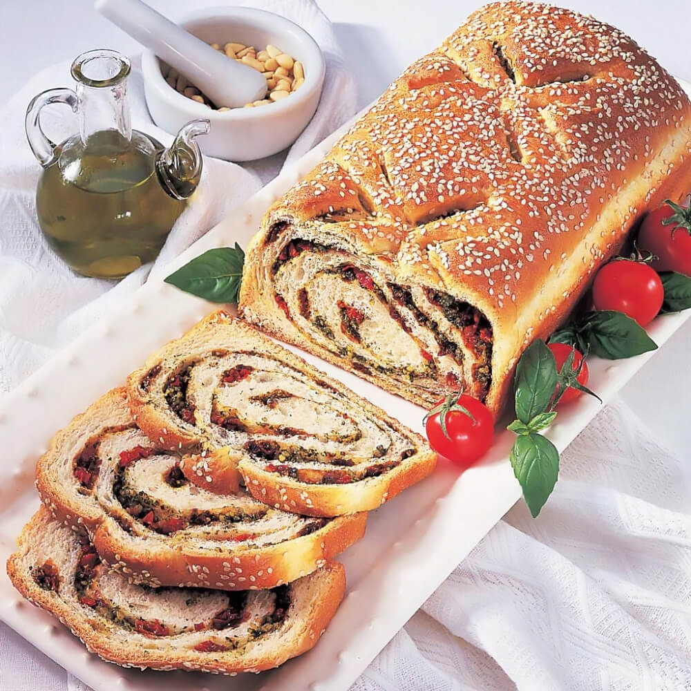 A freshly baked loaf of bread with a golden-brown crust and sesame seed topping sits on a white rectangular plate. The bread is sliced, revealing a swirl of pesto, sun-dried tomatoes, and herbs. Cherry tomatoes and fresh basil leaves surround the loaf. In the background, a glass bottle of olive oil and a white mortar with pine nuts are placed on a white tablecloth.