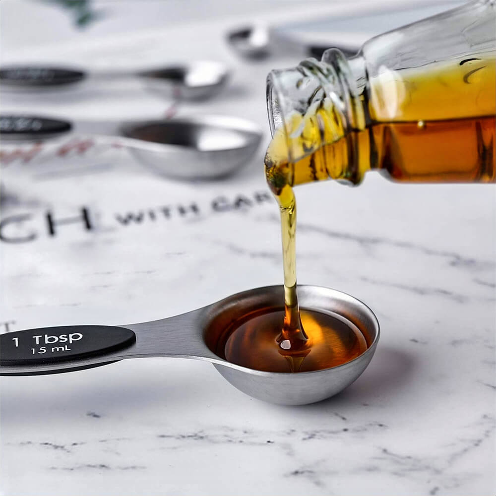 A magnetic measuring spoon filled with syrup being poured from a glass bottle. The stainless steel spoon, labeled "1 Tbsp," is designed for precise measurements.