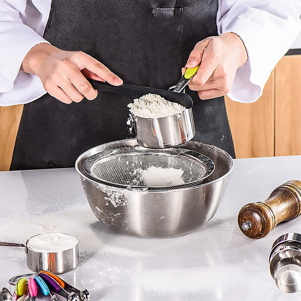 Person using a measuring cups and spoons stainless steel set to sift flour into a metal bowl. The cups have colorful silicone handles and engraved measurements, with additional cups and spoons visible on the counter.
