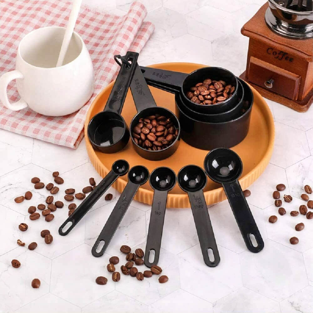 Black measuring spoons and cups filled with coffee beans, placed on a tray with scattered beans on a tiled kitchen counter.