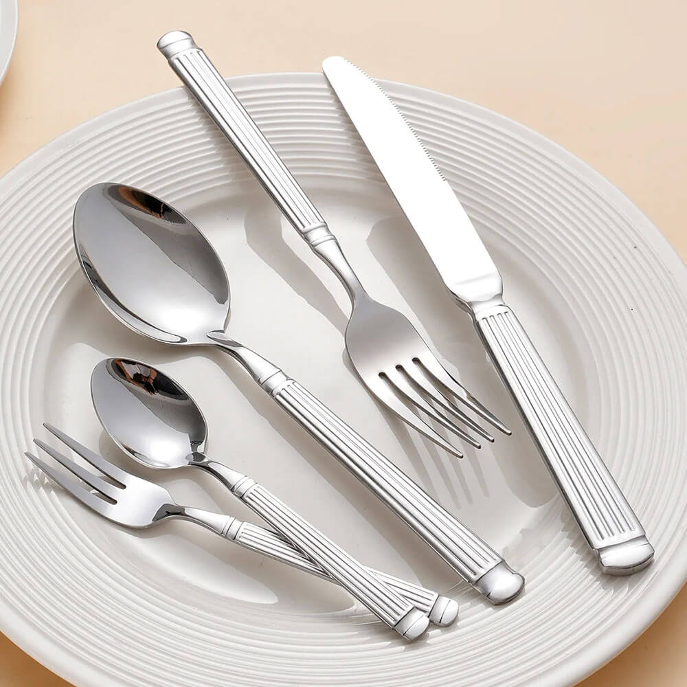 Stainless steel flatware set with grooved handles placed on a white plate, includes forks, spoons, and a serrated knife.