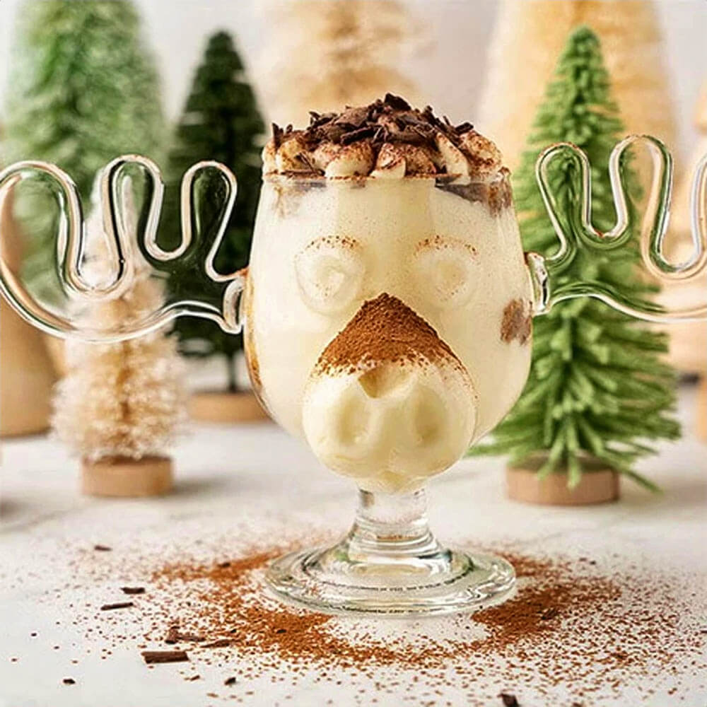 A festive drink is served in a glass from the Moose Mugs Christmas Vacation collection, featuring a moose design with antlers as handles and a sculpted snout. The glass is filled with a creamy beverage, topped with whipped cream, chocolate shavings, and a dusting of cocoa powder. In the background, decorative Christmas trees add a festive touch, emphasizing the holiday theme inspired by the movie National Lampoon&