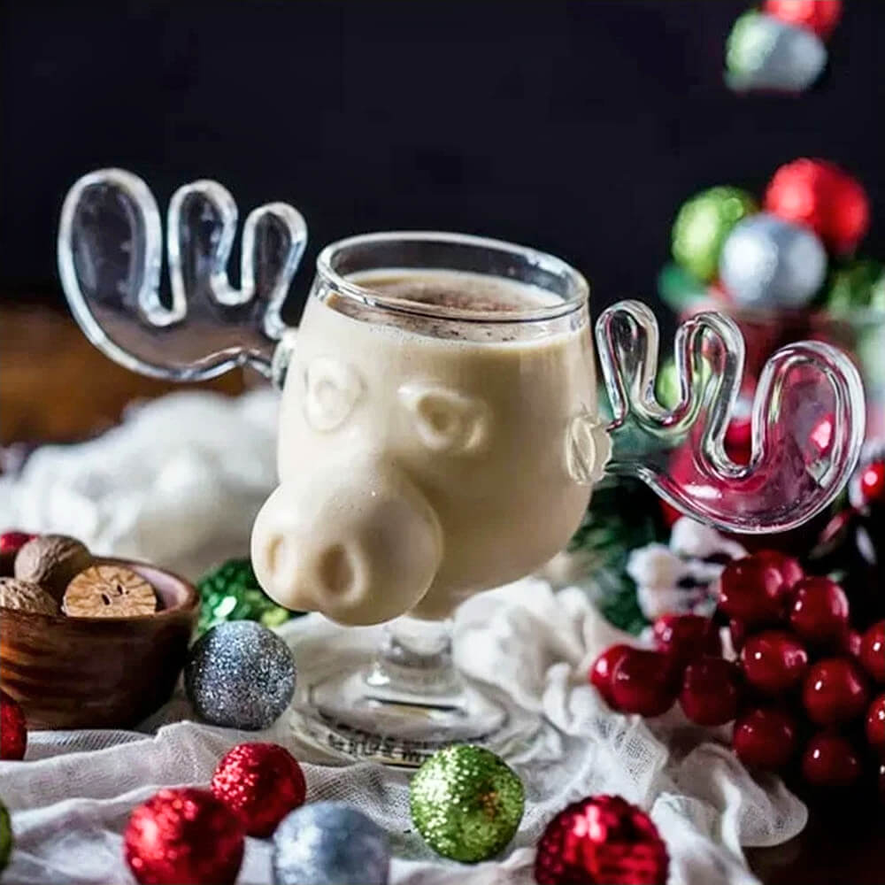 A glass from the National Lampoon Moose Mugs collection filled with a creamy beverage, likely eggnog, topped with a sprinkle of nutmeg. The glass features a detailed moose design with sculpted antlers serving as handles and a prominent moose snout. Surrounding the mug are festive decorations, including colorful glittery ornaments in red, green, and silver, a wooden bowl with whole nutmeg, and a soft white fabric creating a cozy holiday setting. In the background, blurred red and green holiday decorations ad