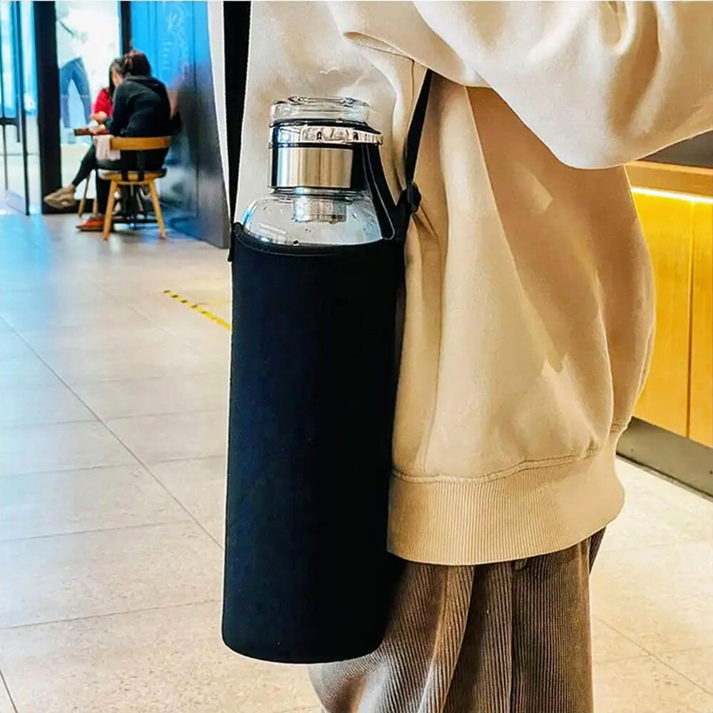 A glass sports water bottle secured in a black neoprene bottle sleeve with a shoulder strap, worn crossbody by a person in a beige hoodie. The sleeve provides added protection and portability, making it easy to carry the bottle hands-free. The background features a modern café setting with seating and warm lighting.