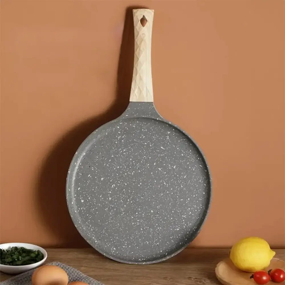 Gray crepe pan with a light wooden handle placed upright against a brown wall, shown with eggs, lemon, and vegetables on a table.