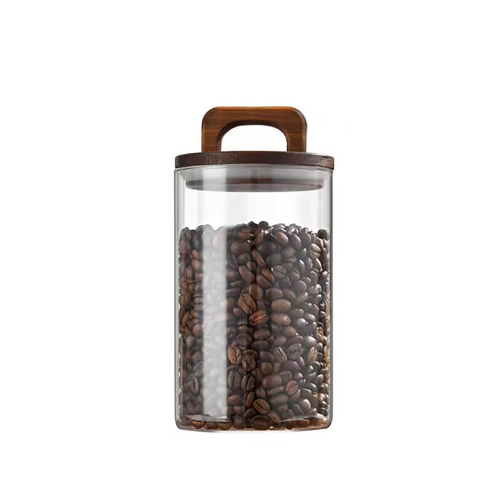 A transparent pantry glass jar filled with coffee beans, featuring a cylindrical shape and a dark wooden lid with a built-in handle, set against a plain white background.