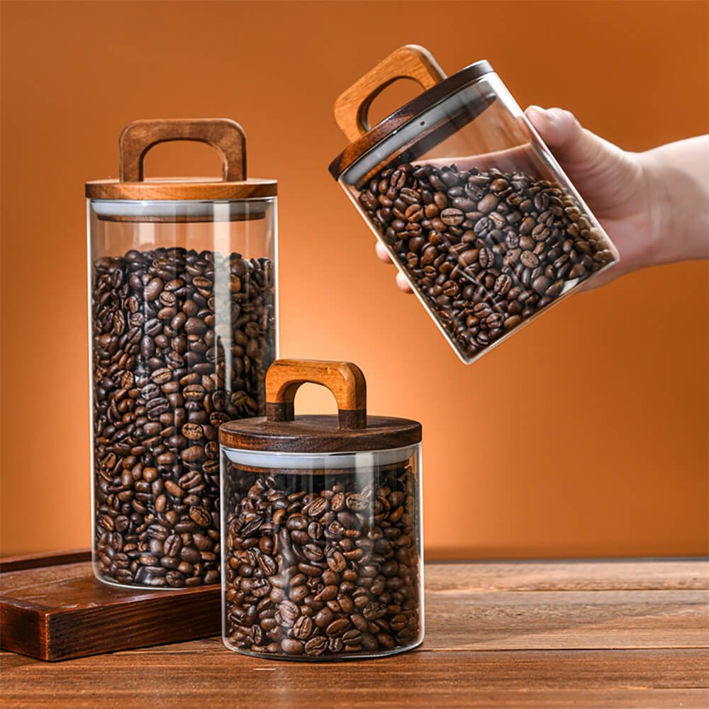 A set of pantry storage jars filled with coffee beans, featuring transparent glass bodies and wooden lids with built-in handles. One jar is being held at an angle by a hand, while the other two are placed on a wooden surface against a warm-toned background. The jars vary in size and have an airtight seal for freshness.