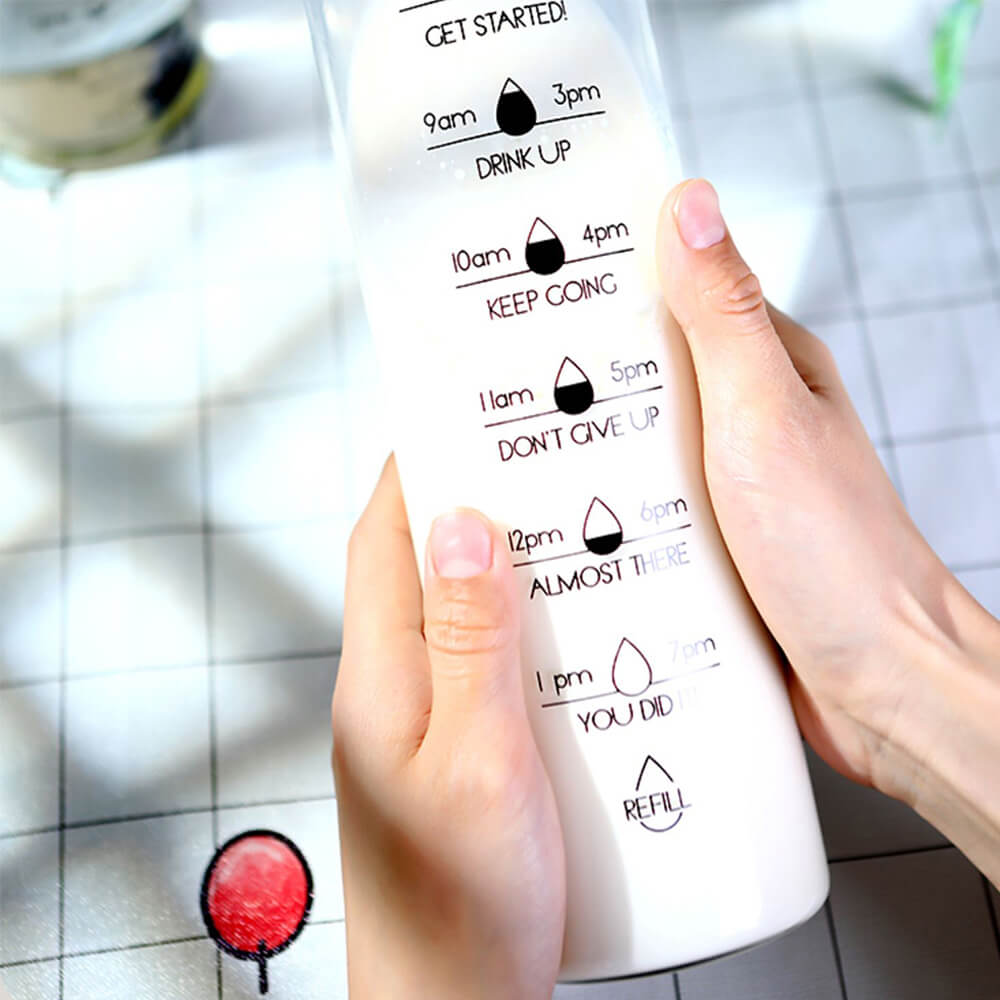 A person holding a reusable glass bottle with a white liquid inside featuring time markers from 8 AM to 7 PM and motivational phrases like KEEP GOING, DON’T GIVE UP, and REFILL to support regular hydration throughout the day.