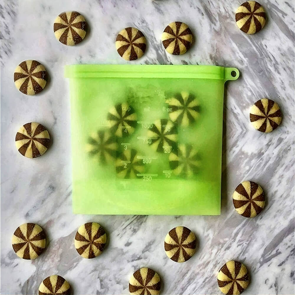 Green reusable silicone bag filled with pinwheel cookies, placed on a marble countertop and surrounded by matching cookies arranged in a circle.