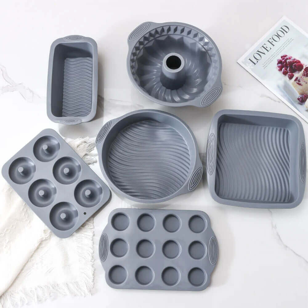 Set of six gray silicone bakeware molds including muffin, donut, loaf, round, bundt, and square pans on a white surface.