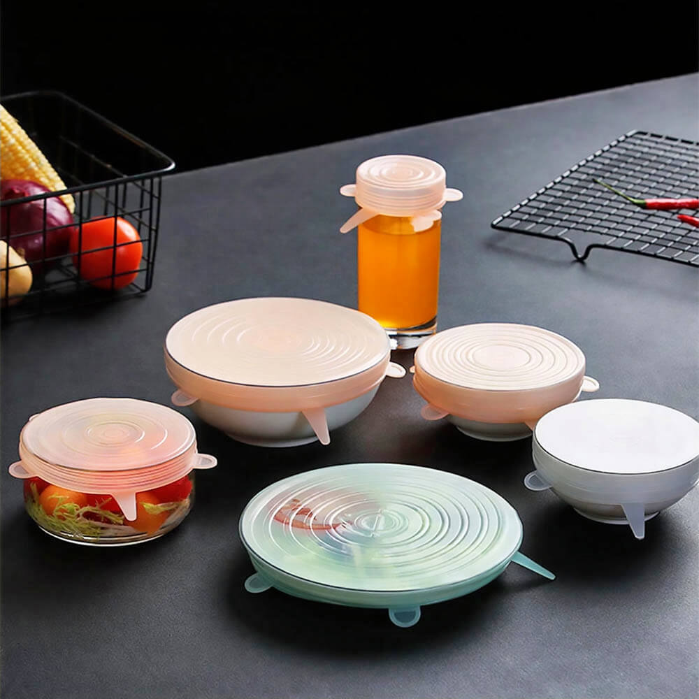 Six assorted containers and a bottle covered with round, translucent silicone stretch lids on a dark kitchen counter.