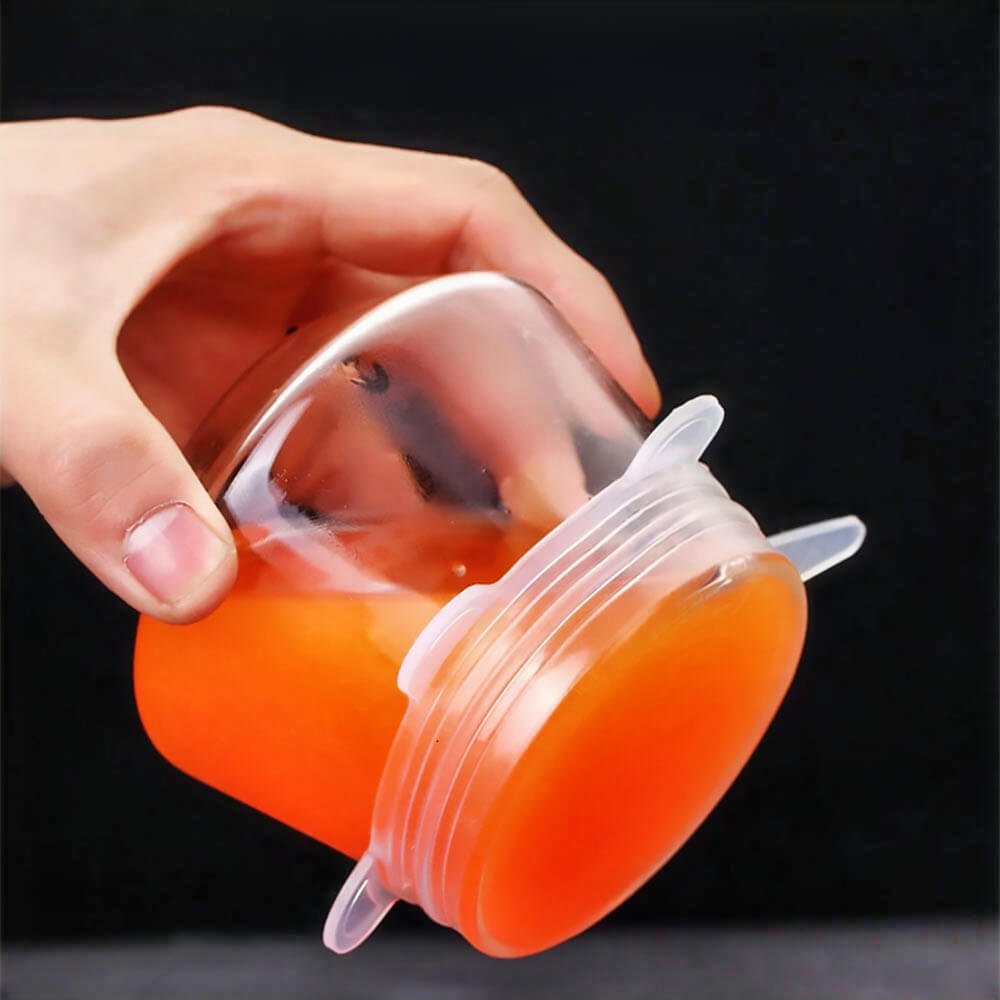 Hand holding a glass cup sealed with a clear silicone stretch lid, securely containing orange liquid without spills.