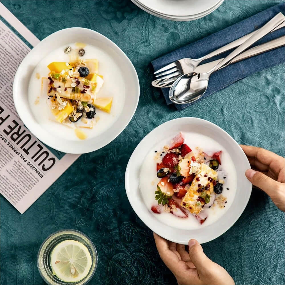 Two white snack bowls filled with creamy fruit desserts, featuring fresh berries, mango, and granola, placed on a teal tablecloth with cutlery and a lemon drink nearby.