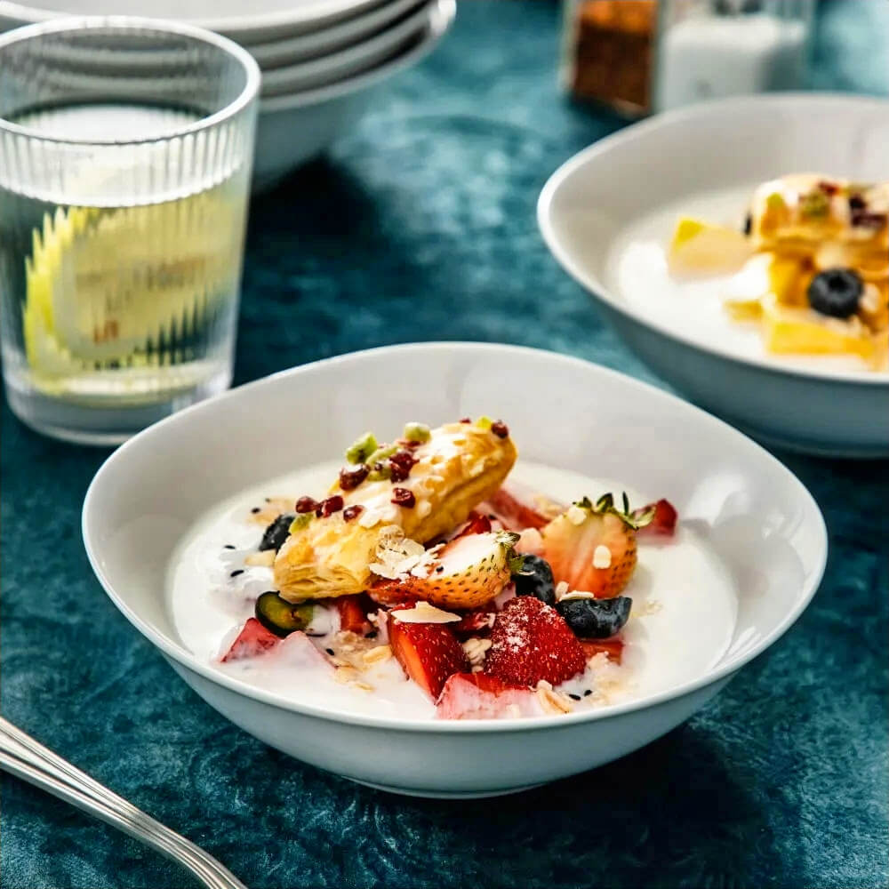 White ceramic snacking bowls filled with creamy fruit desserts, including strawberries, blueberries, and pastry, placed on a dark teal table with a glass of lemon water and stacked bowls in the background.