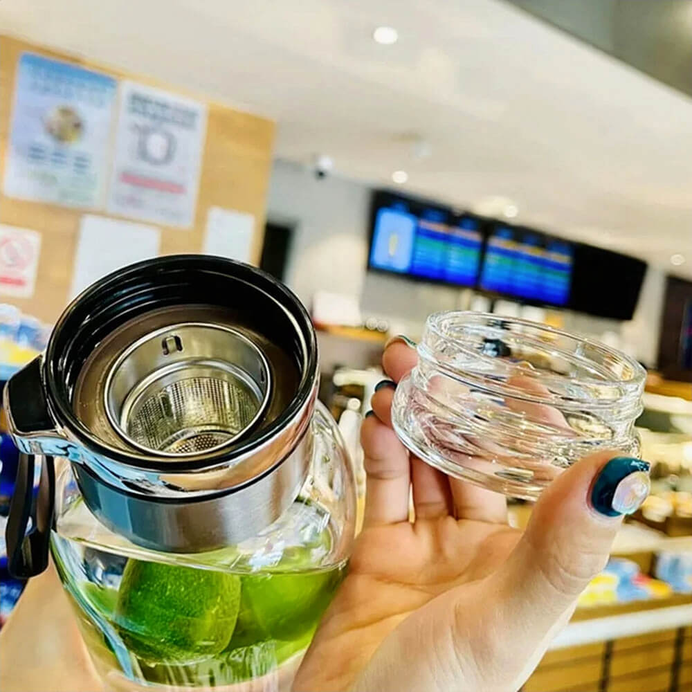 A glass sports water bottle with a stainless steel leakproof lid and a built-in metal filter, being held by a person. The bottle contains water with green limes, showcasing its use for infused drinks. The wide-mouth opening and detachable lid allow for easy filling and cleaning. The background features a modern café setting.