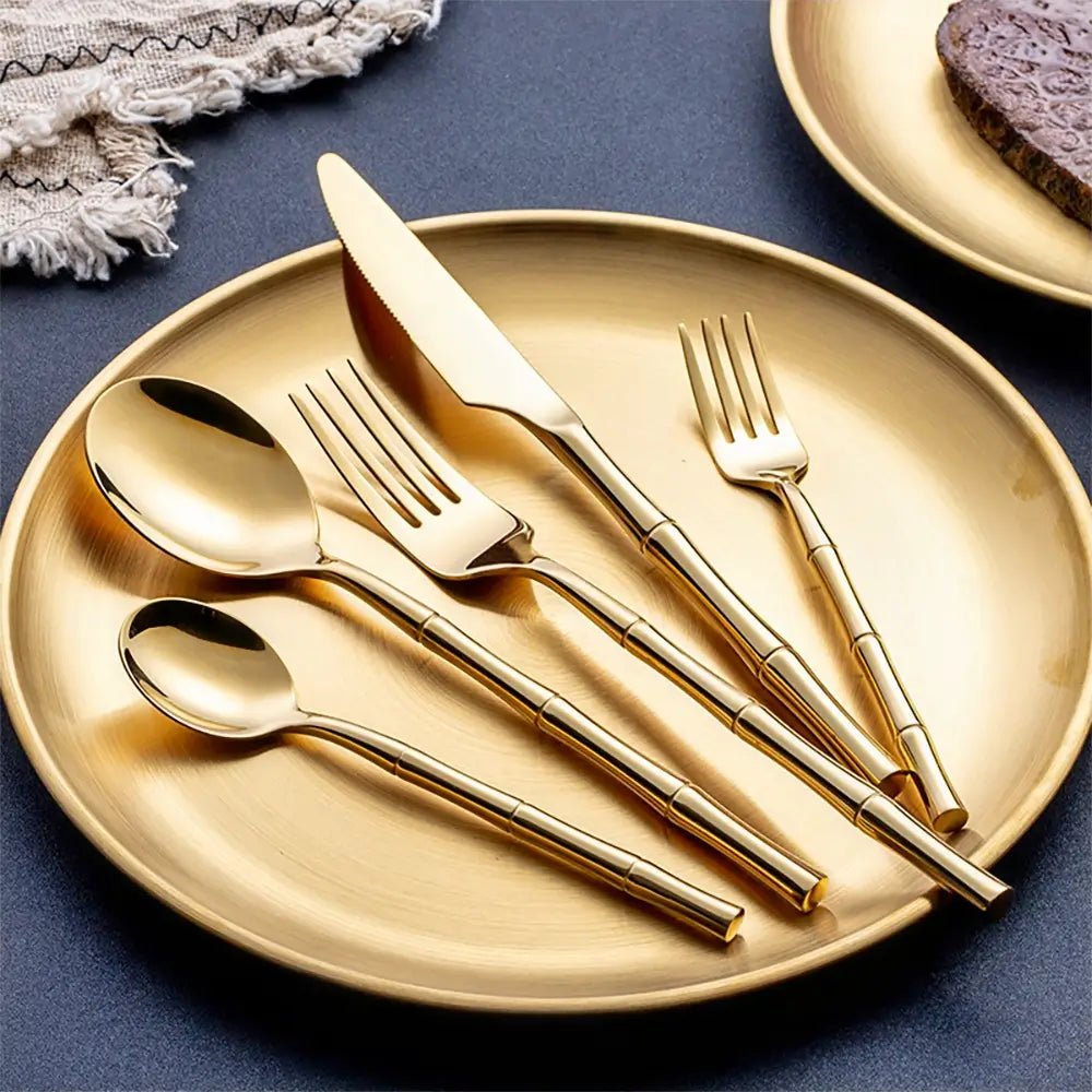 Gold stainless flatware 18 10 set arranged on a matching gold plate with bamboo inspired handles on a dark tabletop.