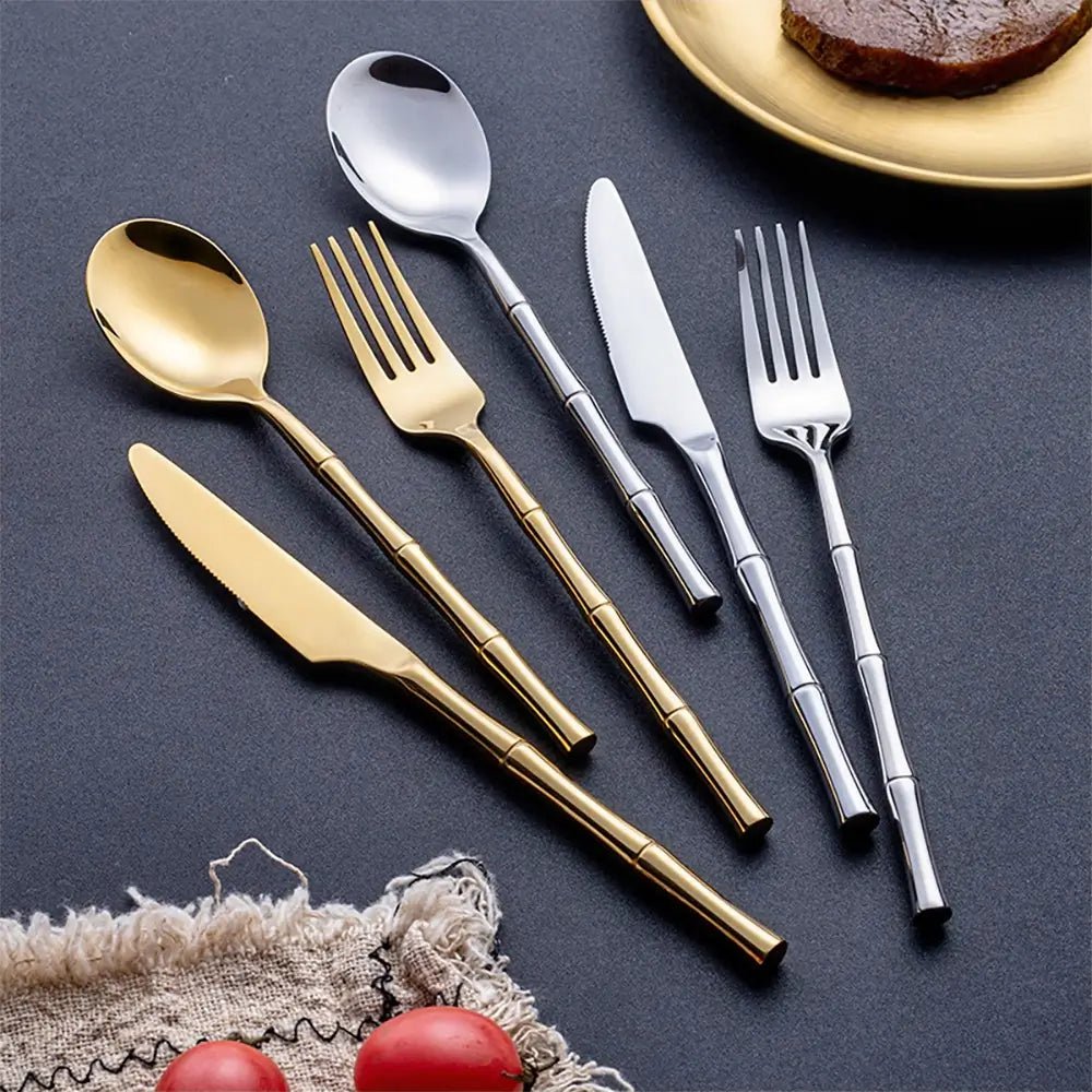 Modern stainless steel flatware set in silver and gold with sleek bamboo-style handles, arranged on a dark tabletop.