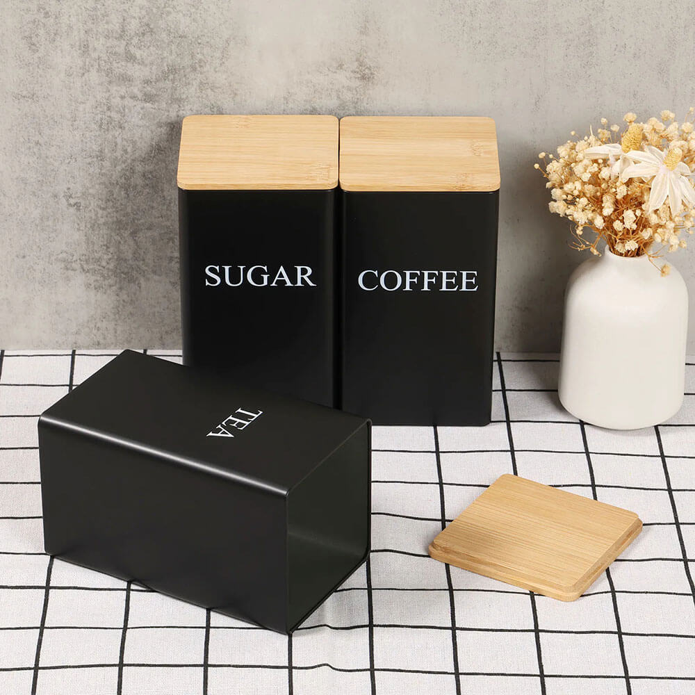 Three black stainless steel food storage containers labeled SUGAR, COFFEE, and TEA with bamboo lids placed on a checkered cloth.