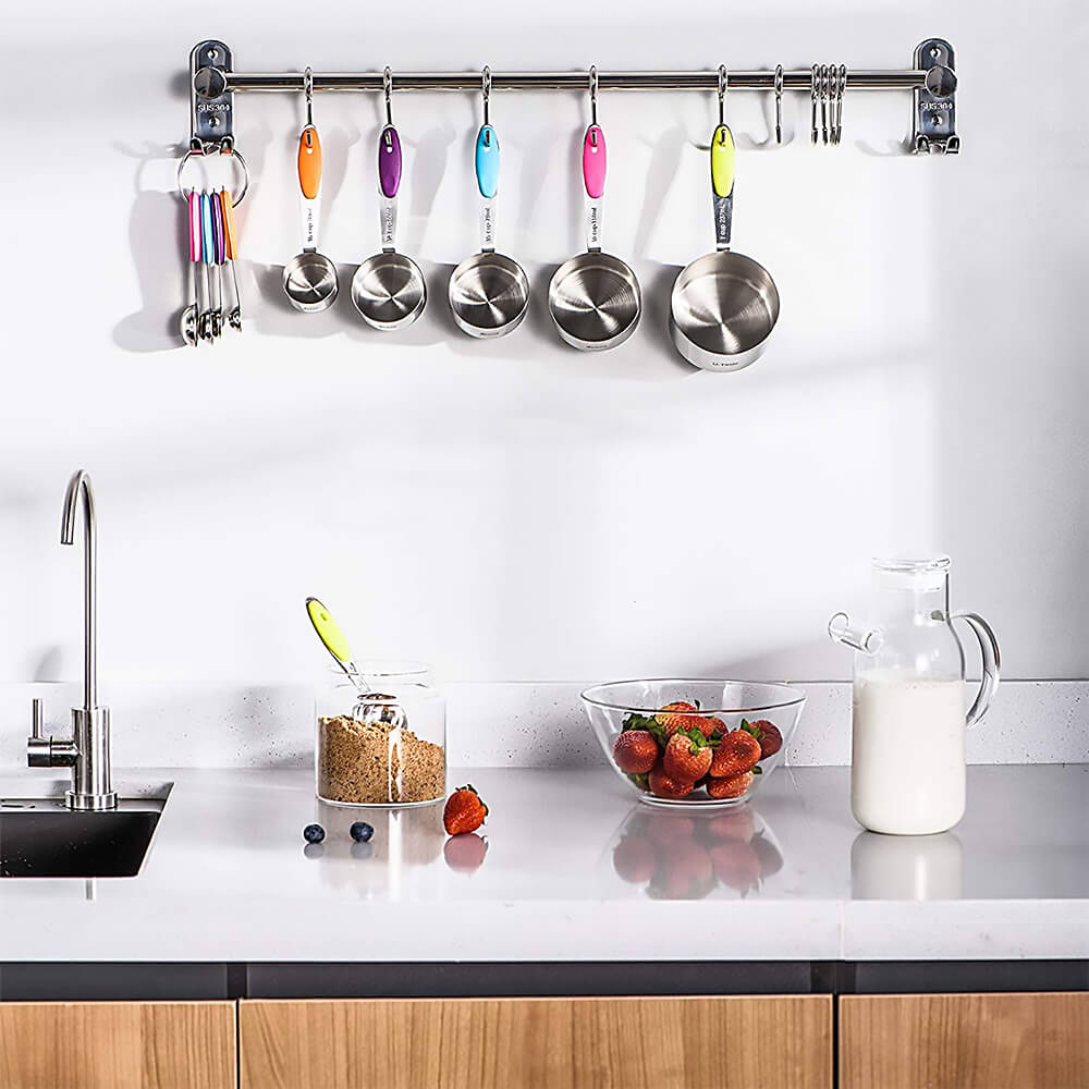 Stainless steel measuring cups and spoons with colorful silicone handles neatly hanging on a kitchen wall rack above a countertop with strawberries, brown sugar, and a glass jug of milk.
