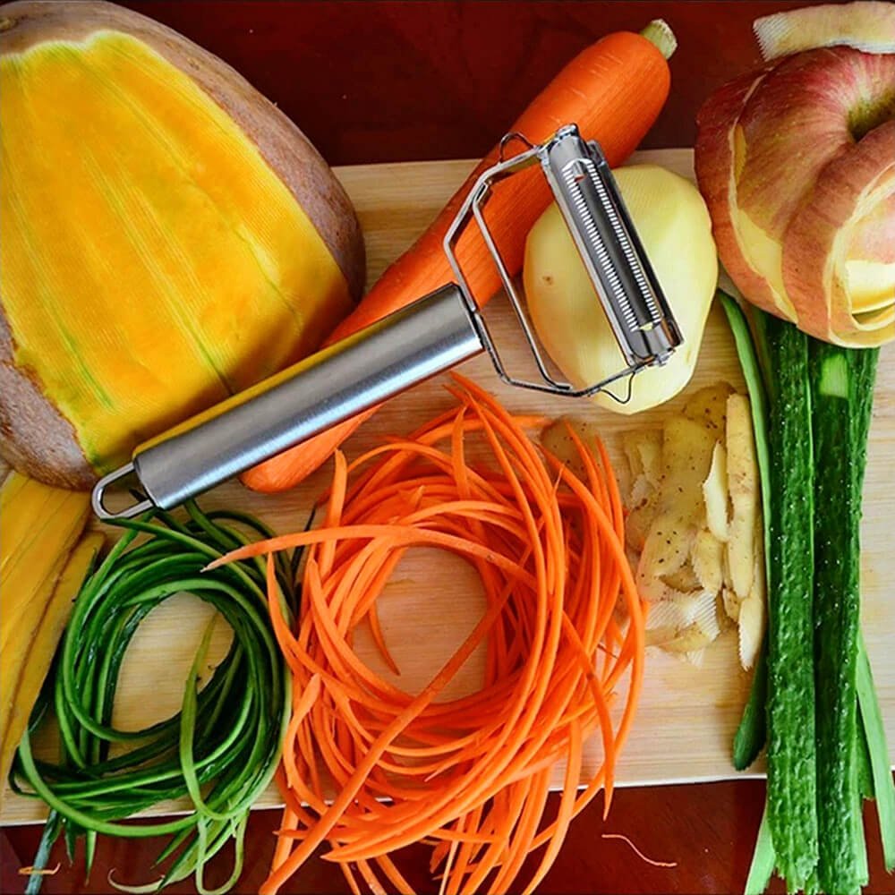 Stainless steel vegetable peeler on a wooden cutting board, surrounded by peeled and julienned carrots, cucumbers, potatoes, and apples. Ideal for precise food preparation.