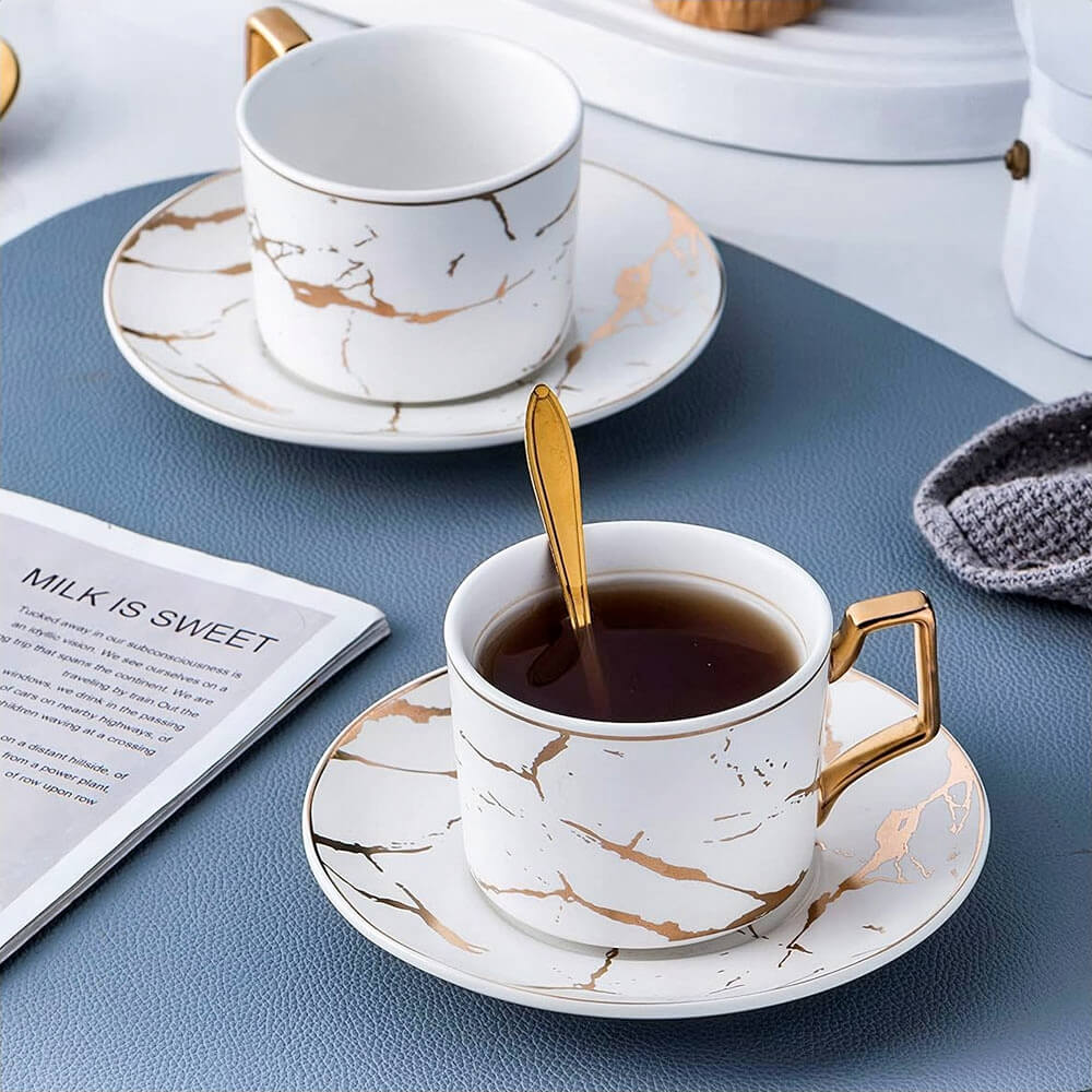 A beautifully arranged tea setup with two teacups filled with tea, resting on matching saucers with a marble design and gold accents. One golden spoon is placed inside a cup, while a magazine titled "MILK IS SWEET" and a textured blue mat add to the cozy, refined ambiance.