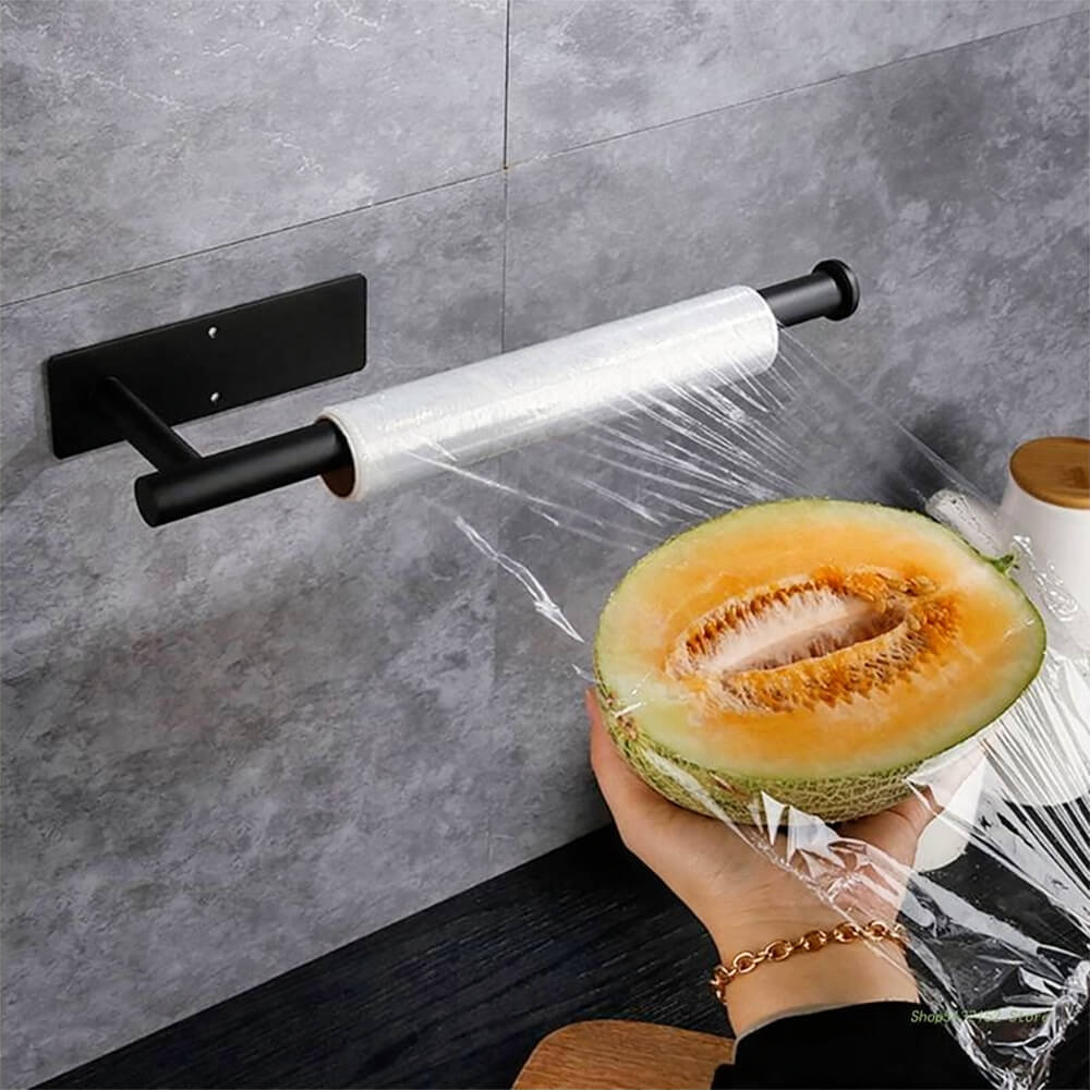 Wall mount paper towel holder used to dispense plastic wrap over a halved melon on a kitchen counter with gray backsplash.