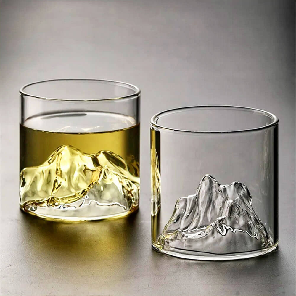 Two whiskey glasses with mountains are displayed, each featuring a sculpted mountain peak design at the base. One glass is filled with a golden liquid, showcasing how the beverage accentuates the intricate details of the mountain design, while the other glass remains empty, highlighting the clarity and craftsmanship of the glass.