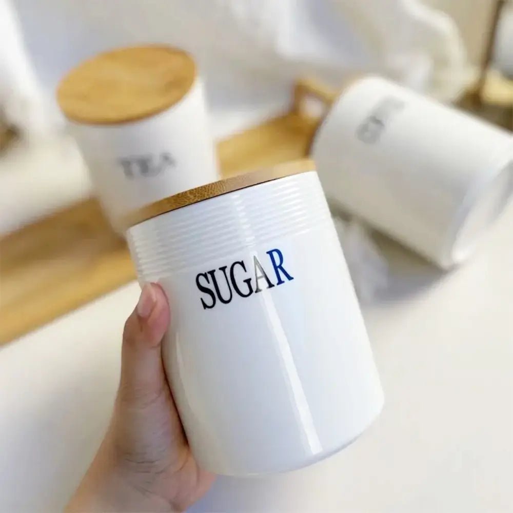 White tea coffee sugar canisters with bamboo lids, one sugar canister held in hand, others on a wooden tray in background.