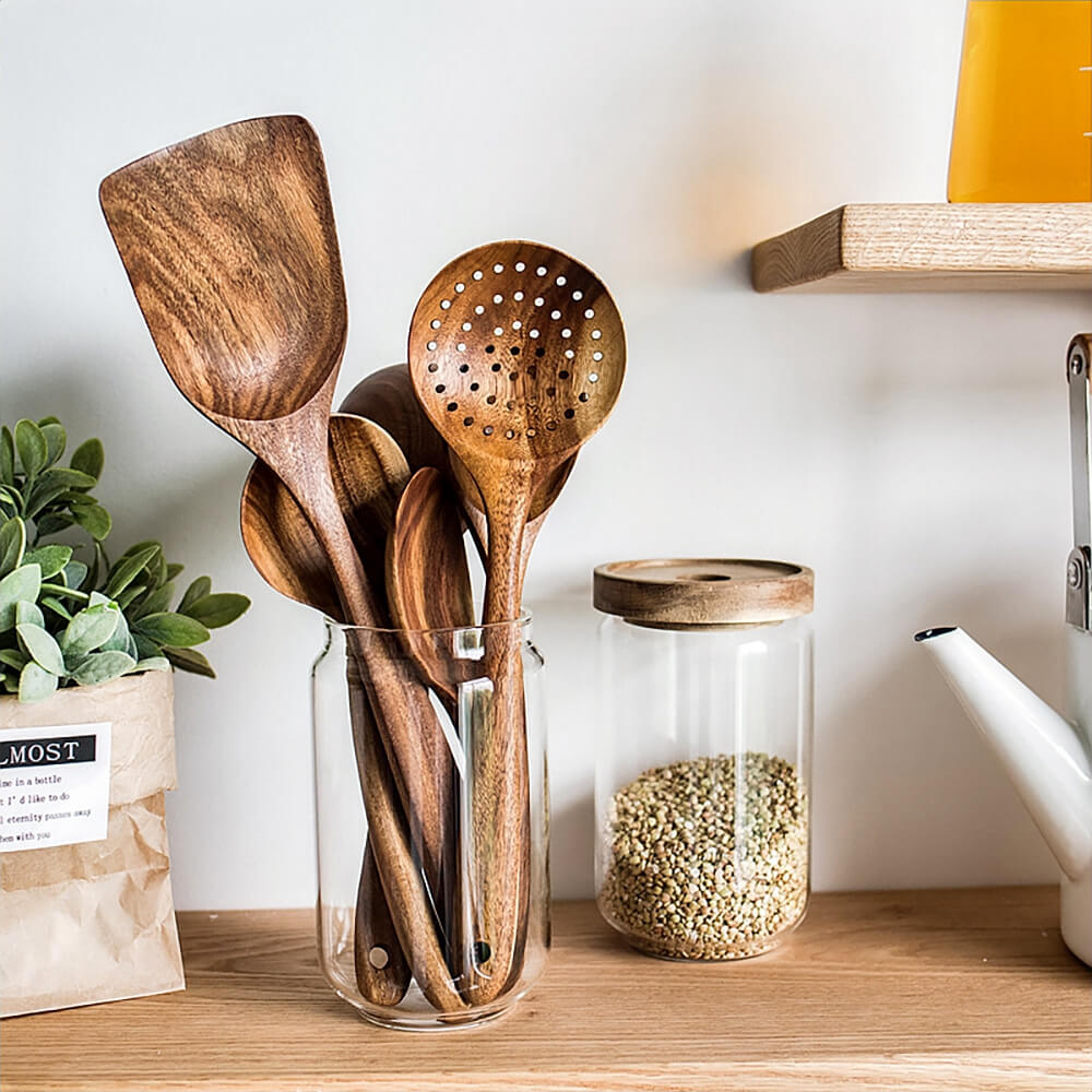 Wooden cooking utensil set displayed in a glass jar on kitchen shelf, includes spatulas, ladles, and slotted spoon.