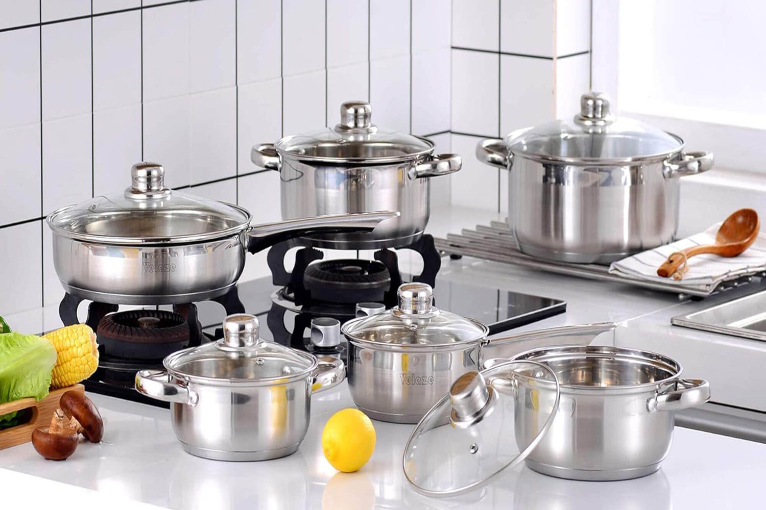 A modern kitchen with a set of stainless steel cookware, including pots and pans with glass lids, arranged on a gas stove and countertop. The cookware set has a polished silver finish, reflecting the bright, clean kitchen environment with white tiled wall