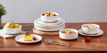 Elegant white dinnerware set on wooden table with fruit, salad, and dessert, showcasing modern minimalist design.