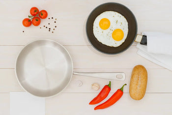 Top view of a stainless steel pan and a non-stick pan with sunny-side-up eggs, surrounded by fresh vegetables and spices.
