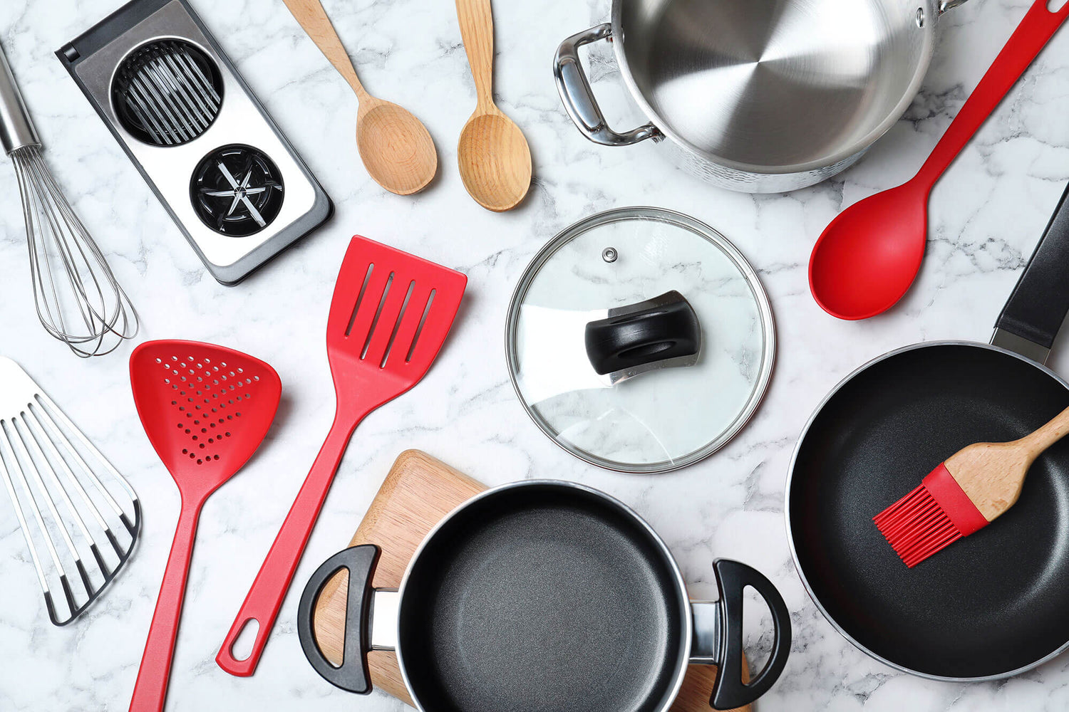 A flat lay of top kitchen gifts, including nonstick frying pans, stainless steel pots, red silicone spatulas, wooden spoons, a whisk, a slicer, and a glass lid, all arranged on a white marble countertop. These essential kitchen items for gifting combine s