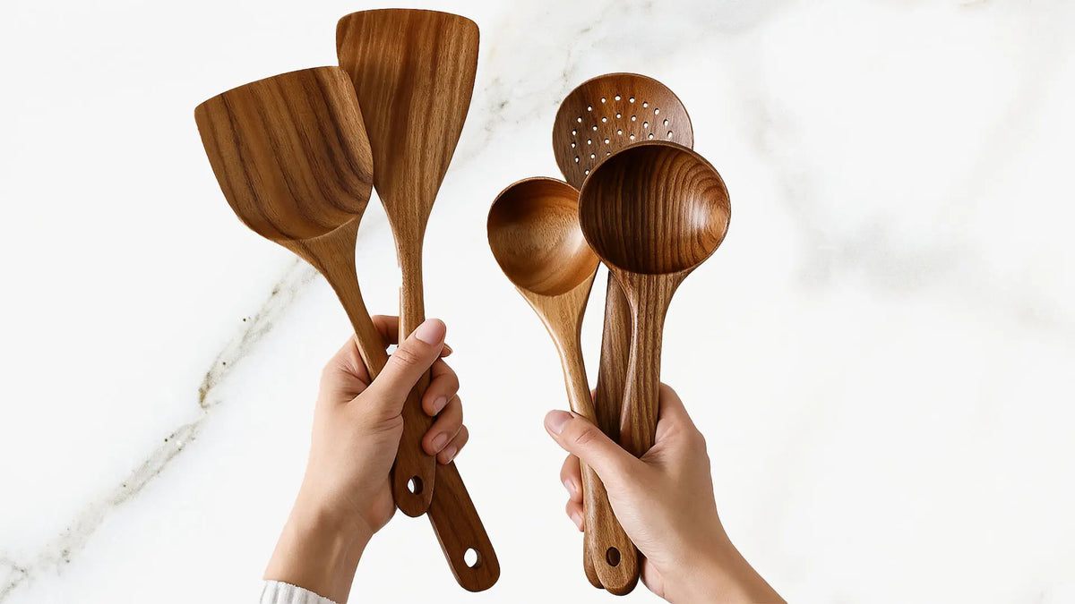 Two hands holding a variety of wooden cooking utensils, including spatulas and ladles, against a white marble background.