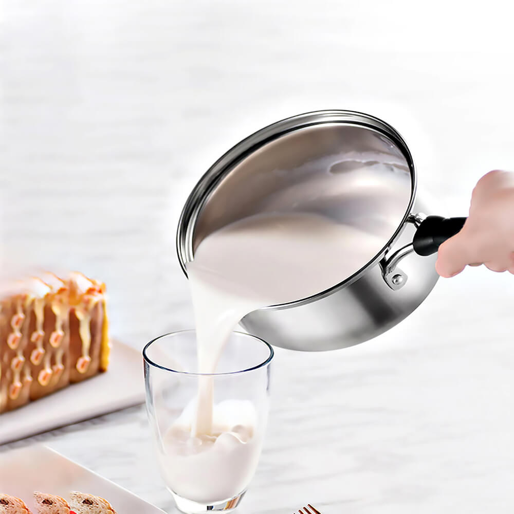 Hand pouring milk from a 1 quart stainless steel saucepan with lid into a clear glass, with dessert items on a white table.