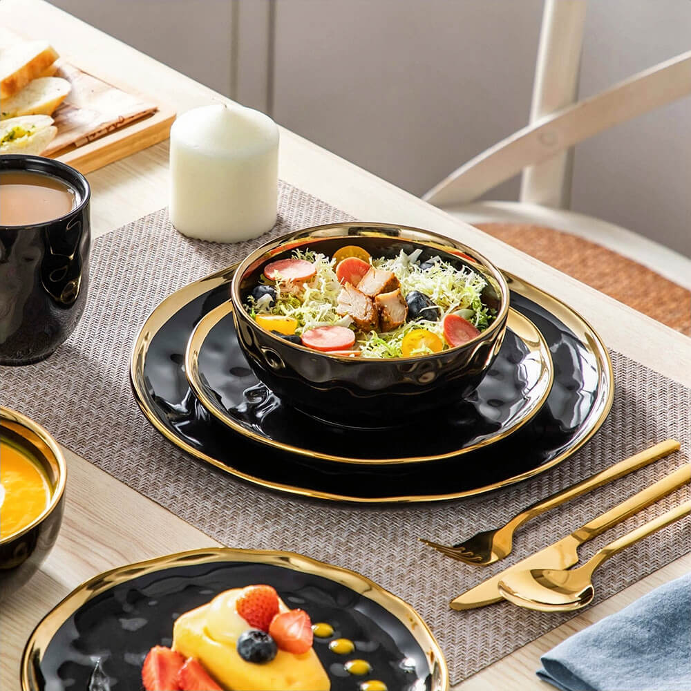 A 16-piece stoneware dinnerware set arranged on a dining table. The set features glossy black plates, bowls, and mugs with gold accents on the rims and edges. A bowl is filled with a vibrant salad of greens, cherry tomatoes, blueberries, and grilled chicken, placed on a layered arrangement of plates. A dessert plate displays a dish garnished with fresh strawberries and blueberries. Gold cutlery is neatly placed beside the dinnerware on a beige woven placemat, accompanied by a lit white candle for a cozy din