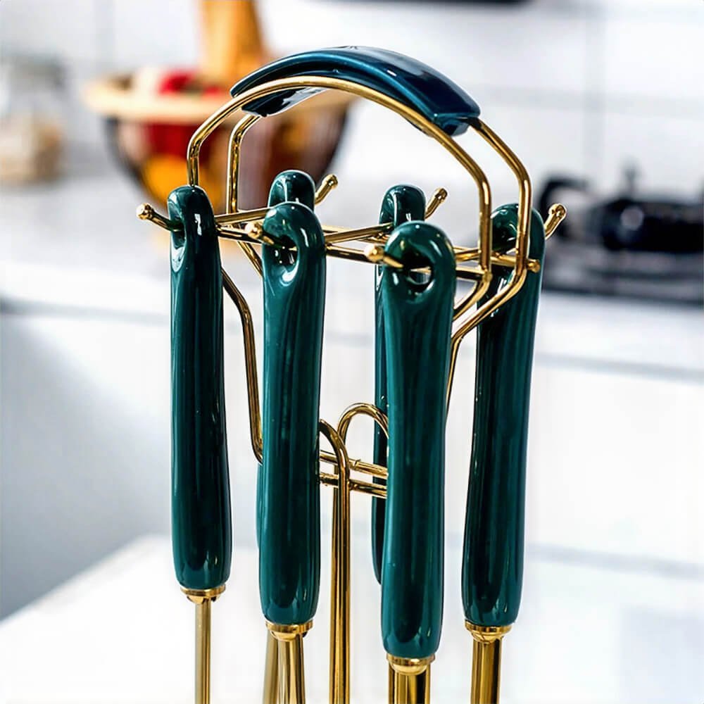 A close-up image showcasing the handles of an 18/0 stainless steel utensil set, featuring glossy dark teal finishes and gold accents. The utensils are neatly arranged on a matching gold stand with a curved handle for easy portability. The blurred kitchen setup in the background adds a modern and stylish context to the scene.