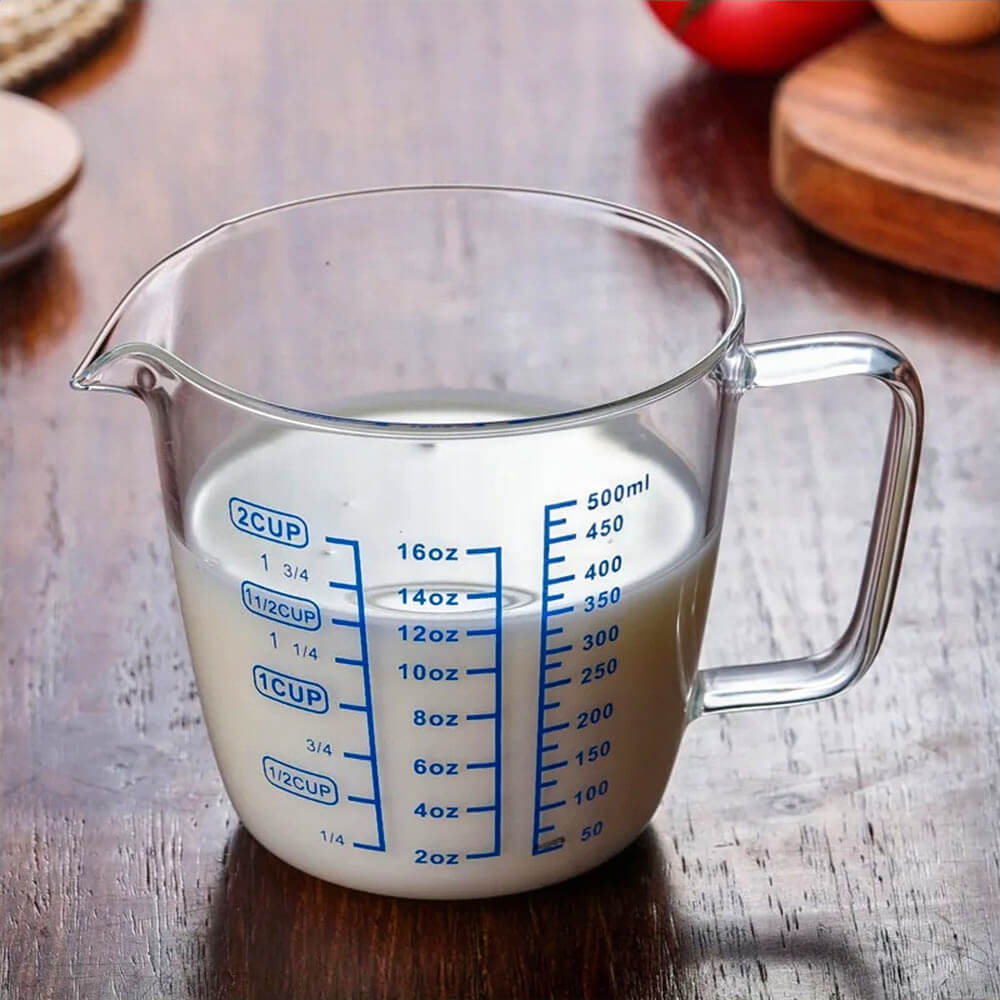 A 2-cup glass measuring cup filled with milk, featuring blue measurement markings for ounces, cups, and milliliters. It has a curved spout and a sturdy handle, placed on a wooden surface.