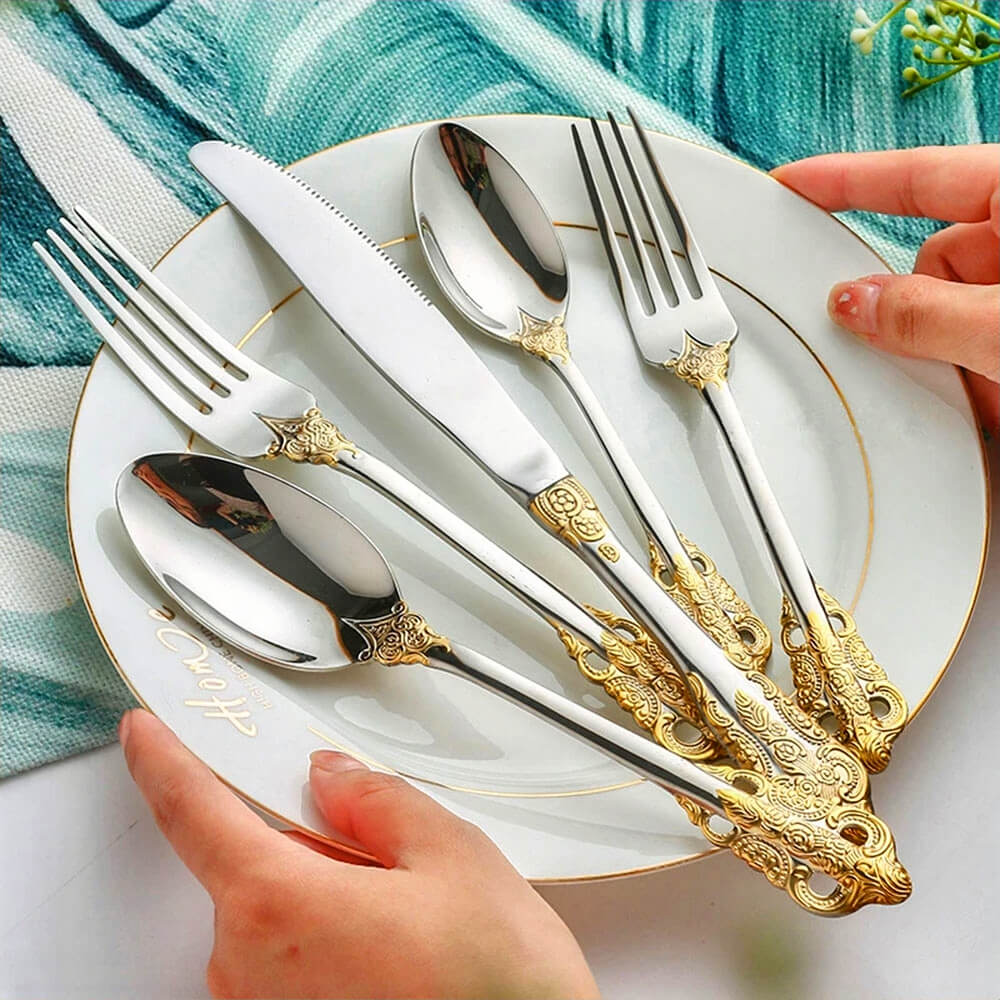 20-piece stainless steel flatware set with ornate gold handles displayed on a white plate with gold rim, held by two hands.