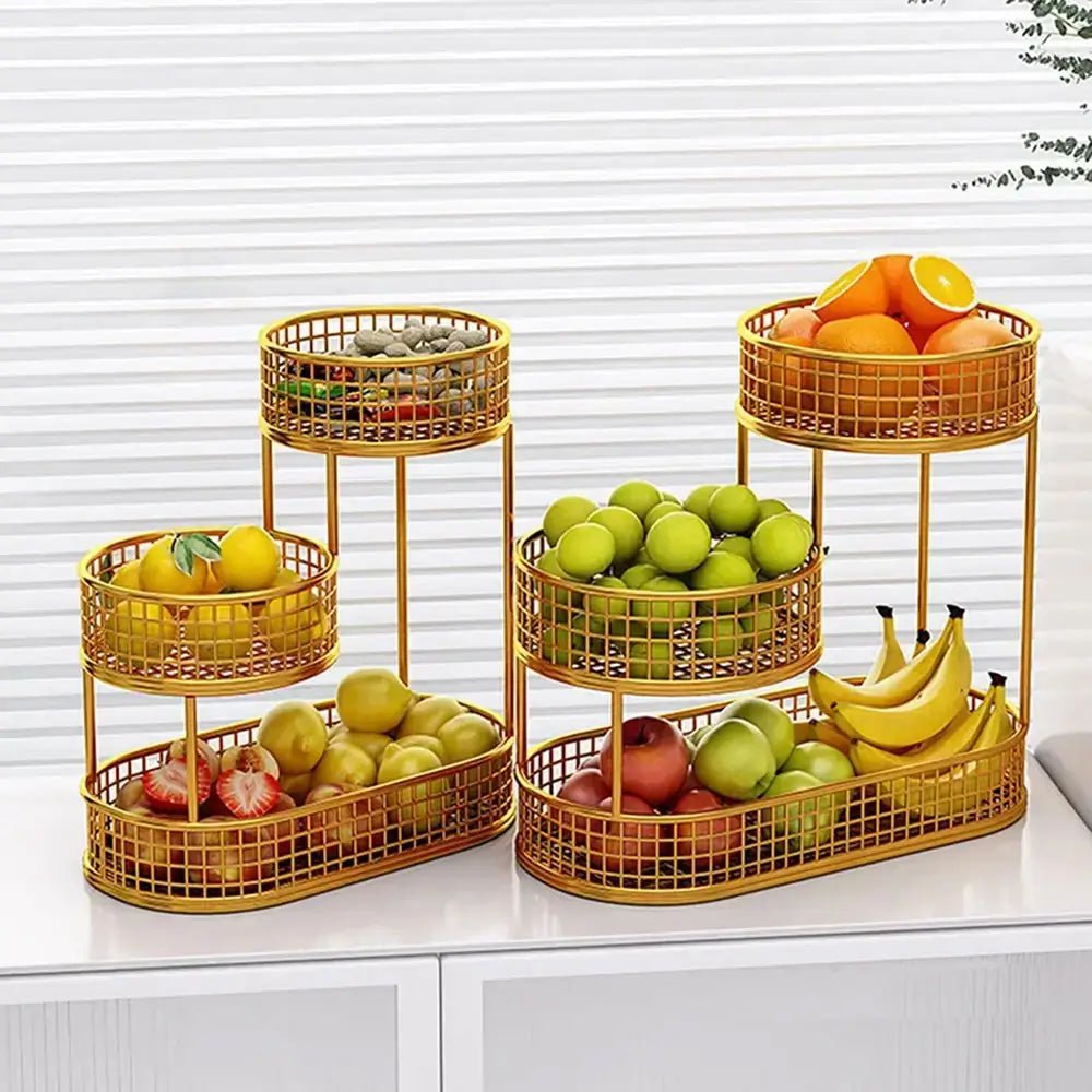 Gold 3 tier countertop fruit basket holding oranges, green apples, bananas, lemons, and strawberries on a white cabinet near a window.