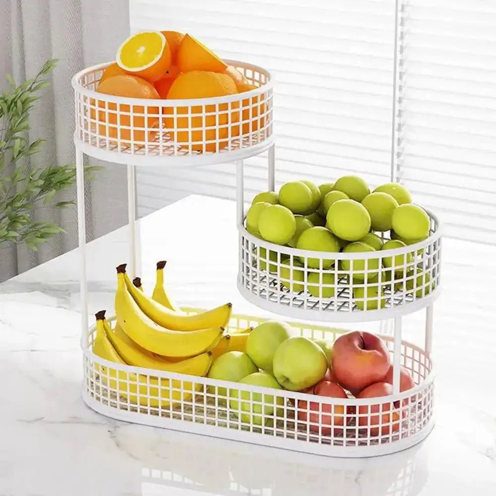 White 3 tier metal fruit basket holding oranges, green apples, bananas, and red apples on a marble countertop near a window.