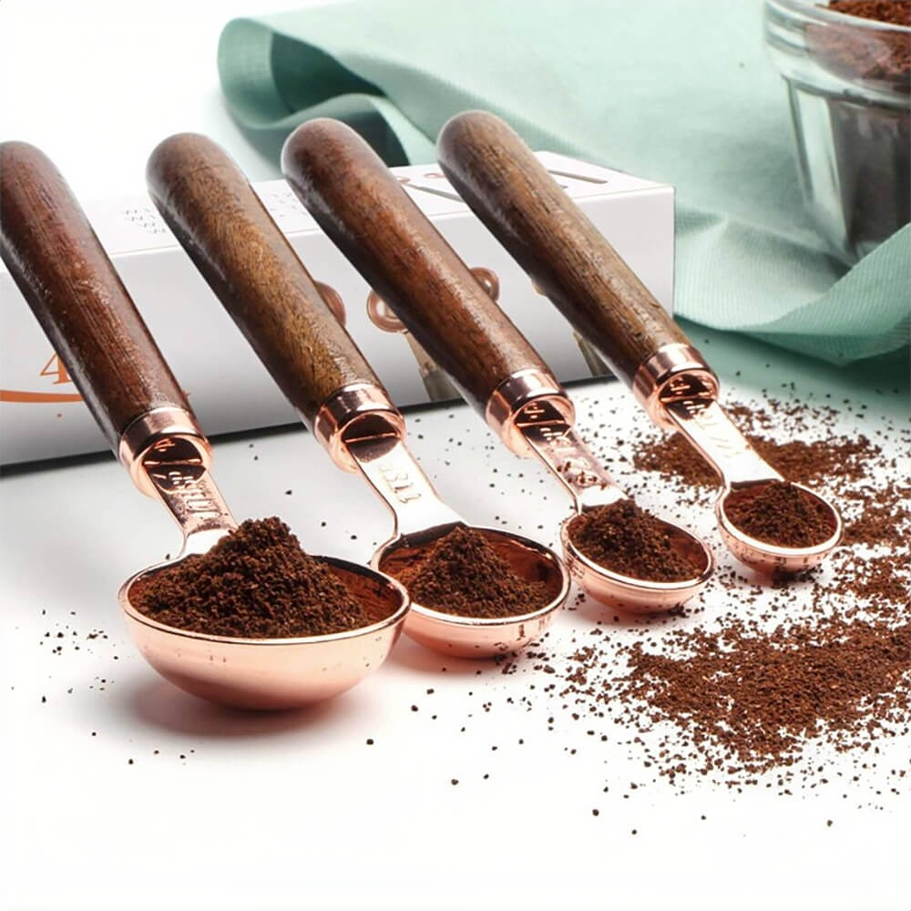 A set of 300mg stainless measuring spoons featuring polished copper-plated heads and dark walnut handles. Each spoon is filled with ground coffee, with scattered coffee grounds adding a rustic touch. A mint green cloth and a glass container create a cozy, modern kitchen backdrop.