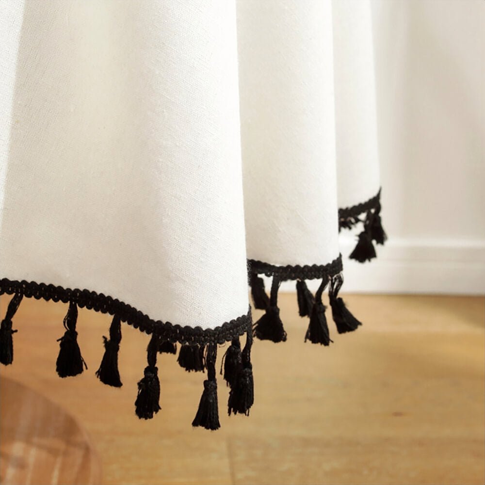 A close-up view of a 90" round tassel tablecloth in white, showcasing its elegant design. The edge of the tablecloth features intricate black trimming and evenly spaced black tassels. The fabric drapes smoothly, creating a clean and sophisticated look suitable for various decor styles.