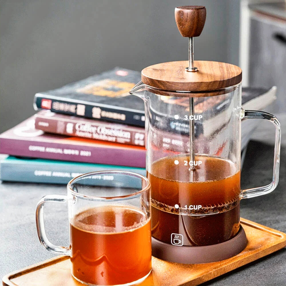 A glass French press with a clear borosilicate glass body, a walnut wood lid, and a stainless steel plunger sits on a wooden tray next to a matching glass mug filled with freshly brewed coffee. The all-glass French press has measurement markings for 1, 2, and 3 cups, a sturdy glass handle, and a brown silicone base for stability. In the background, a stack of coffee-related books adds a cozy, café-style ambiance.