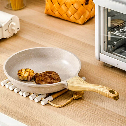 A sleek aluminum fry pan with a natural wood grain handle, showcasing a piece of cooked meat and roasted garlic. The pan sits on a wooden trivet on a light wooden kitchen countertop, near a toaster oven and a basket in a cozy kitchen setting.