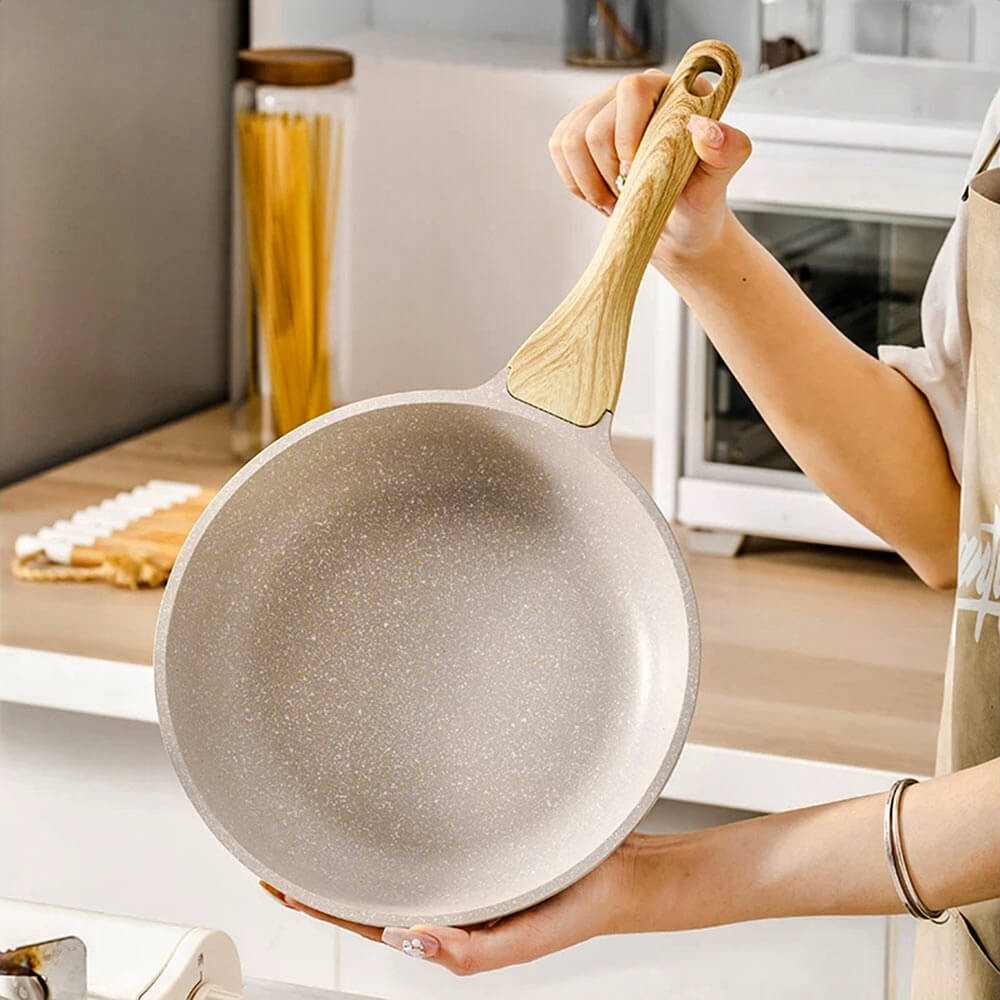Person holding an aluminum frying pan with wood-style handle in a bright, modern kitchen setting.