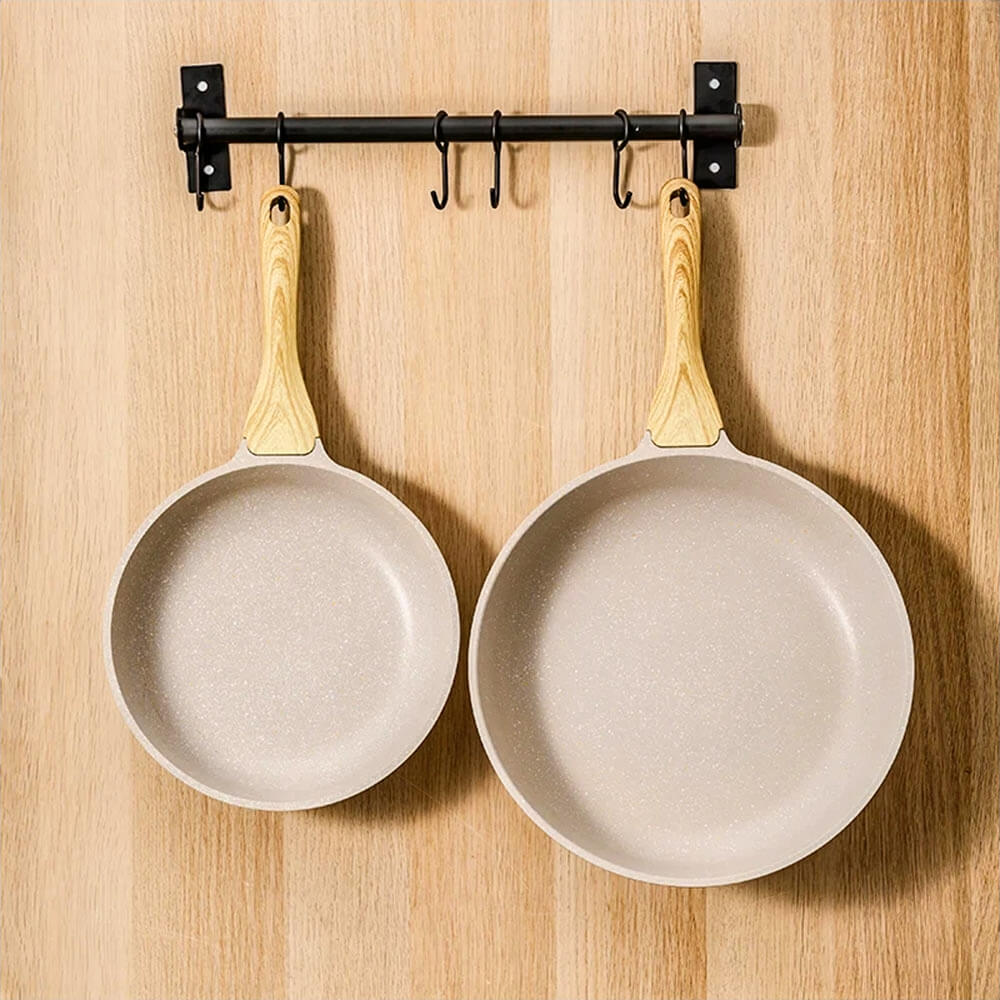 Two aluminum frying pans with natural wood grain handles hanging on a black metal rack against a wooden wall. The pans showcase a smooth, non-stick surface, perfect for modern kitchen organization.