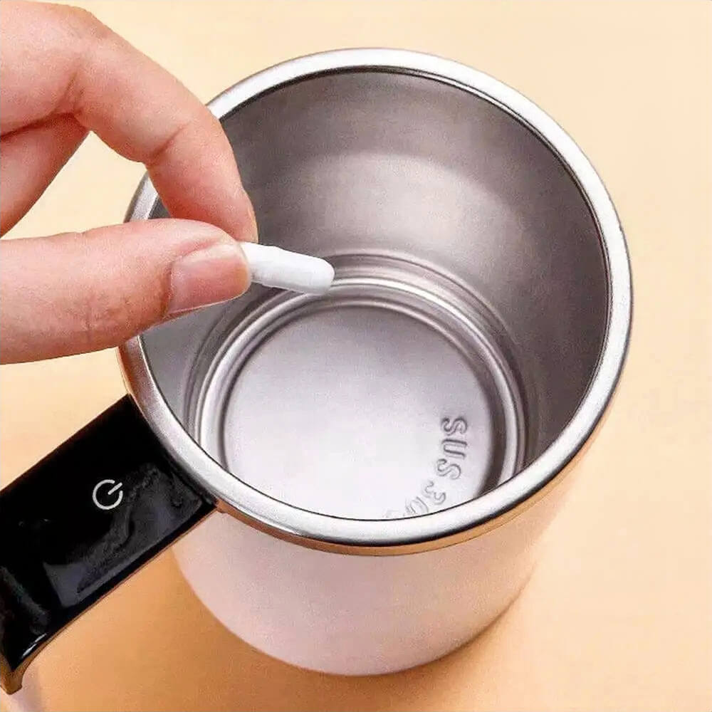 A person is holding a small detachable magnetic stirrer above the interior of an auto coffee mug made from stainless steel. The mug features a polished inner surface and a visible power button on the black handle.