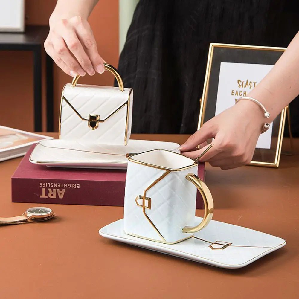 Person holding a bag shaped mug with gold handle on matching saucer, another mug and saucer set placed on a book.